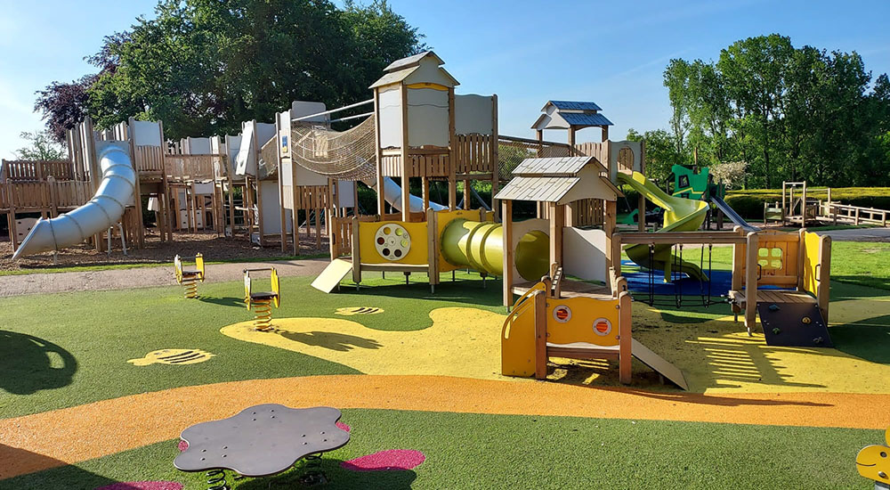 sacrewell outdoor play park in the sunshine. Astro turf with patterned yellow and pink flooring, slides, climbing frames and seasaws.